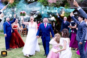 At FF - Espaço de Eventos, guests line the outdoor ceremony path holding vibrant smoke grenades, creating a festive cloud of color as the newlyweds exit, celebrating with infectious joy and excitement.